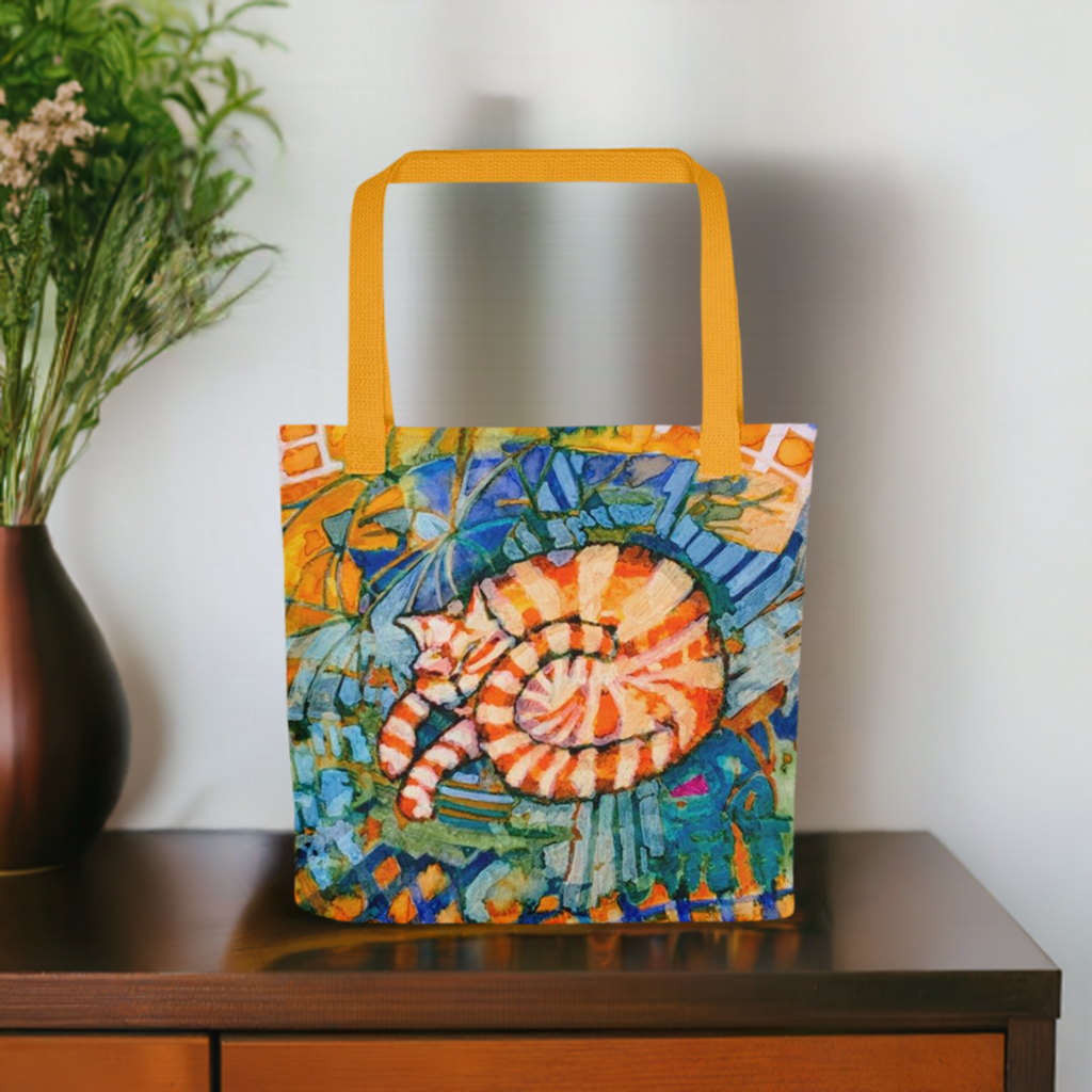 Sleeping Ginger Cat Tote Bag featuring colorful artwork of a curled ginger cat on a vibrant patterned background with yellow handles placed on a wooden surface.