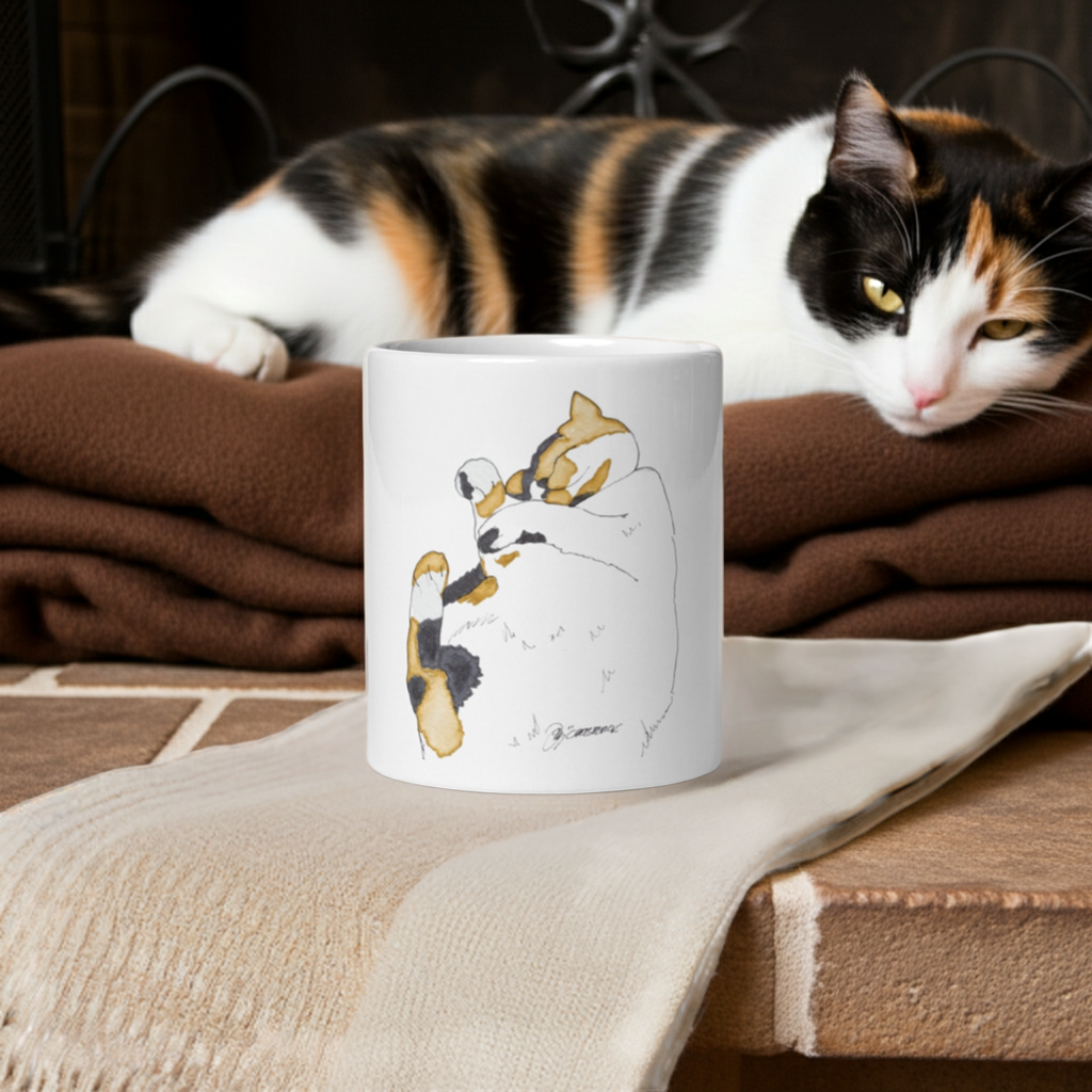 White ceramic napping cat mug with calico cat artwork by Joy Calonico placed on a beige cloth with a calico cat resting in the background